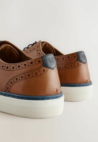 Braune Lederschuhe mit perforierten Details, einem marineblauen Lederakzent an der Ferse und einer weißen, strukturierten Gummisohle.