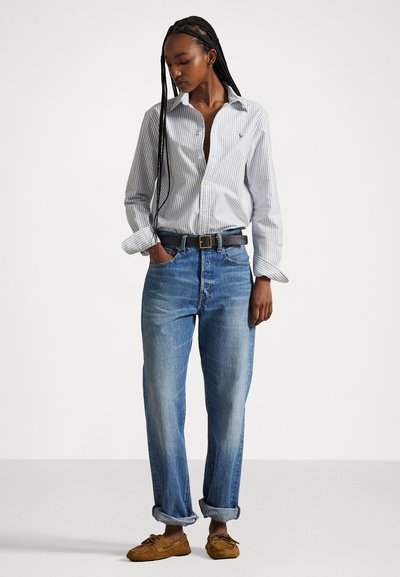 Camisa de botones a rayas blanca y azul, jeans azul relajados, cinturón negro y mocasines marrones. Presenta los puños de los jeans remangados y un logotipo sutil en la camisa.