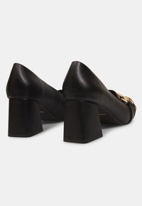 Chaussures à talon bloc en cuir noir avec bouts pointus. Dotées d'un détail en métal doré et de coutures visibles sur les côtés. Texture lisse.