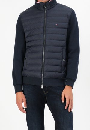 Mann trägt eine dunkelblaue gesteppte Jacke mit Reißverschluss, gerippten Ärmeln und kleinem Logo auf der Brust, kombiniert mit dunklen Jeans.