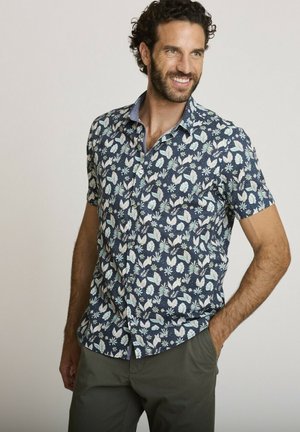 Homme souriant, portant une chemise bleu marine à manches courtes avec un motif floral blanc et un pantalon vert olive, debout avec une main dans la poche.