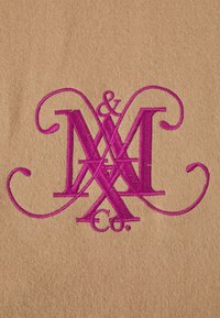 Tissu beige présentant un logo brodé en rose vif avec des lettres ornées, comprenant « M », « & » et « Co. » sur une texture lisse.