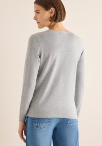 Grå långärmad topp i mjukt tyg, med rund halsringning och avslappnad passform. Kombinerad med blå jeans, visande bakvy och texturerade detaljer.