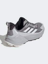 Zapatillas de deporte grises con un upper texturizado, detalles en blanco, una suela de goma gruesa y una lengüeta en el talón. Diseñadas para uso en senderos o al aire libre.