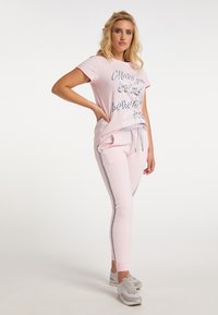 Hellrosa T-Shirt mit grafischem Text, kombiniert mit passenden rosa Jogginghosen mit seitlichen Streifen. Graue Akzente am Bund der Jogginghose und an den Schuhen.