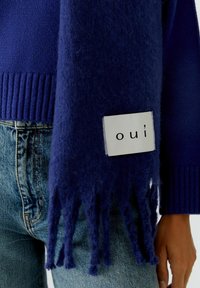 Écharpe bleu marine en matériau doux et pelucheux avec des extrémités à franges et une étiquette blanche portant le texte "oui". Portée avec un jean en denim.