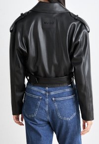 Giacca nera in eco-pelle con spalline sulle spalle, toppa sul retro con logo e dettaglio vita sagomata. Abbinata a jeans in denim.