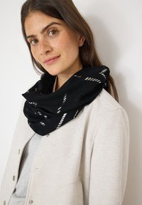 Écharpe noire avec un motif géométrique blanc, fabriquée en tissu léger, portée enroulée autour du cou, accompagnée d'un haut gris clair.