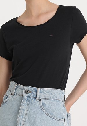 Frau trägt ein schlichtes schwarzes T-Shirt mit kurzen Ärmeln, in hoch taillierte hellblaue Jeans mit Knopf und Gürtelschlaufen gesteckt.