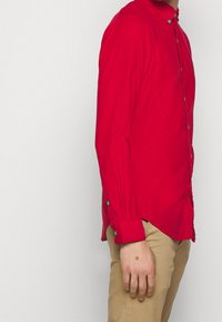 Camicia rossa a maniche lunghe con colletto abbottonato, caratterizzata da bottoni verdi sulla patta e sui polsini. Tessuto liscio con leggero splendore.