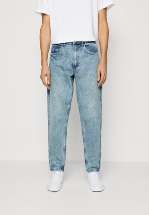 Persoon draagt lichtblauwe jeans met azijnwassing, een eenvoudig wit T-shirt en witte sneakers, staand tegen een witte achtergrond.