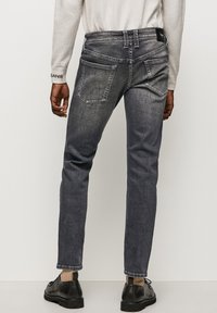 Vaqueros gris oscuro de corte slim, con cinco bolsillos, costuras visibles y un acabado sutilmente texturizado. Llevados con zapatos negros y calcetines tobilleros.