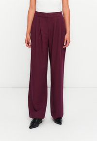 Pantaloni a gamba larga bordeaux con pieghe frontali, vita alta e una texture liscia, abbinati a scarpe nere a punta.