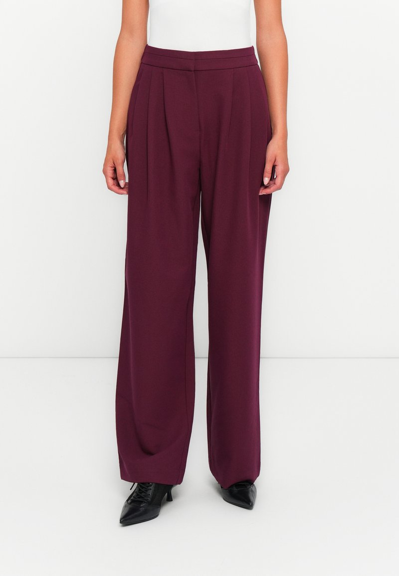 Pantaloni a gamba larga bordeaux con pieghe frontali, vita alta e una texture liscia, abbinati a scarpe nere a punta.