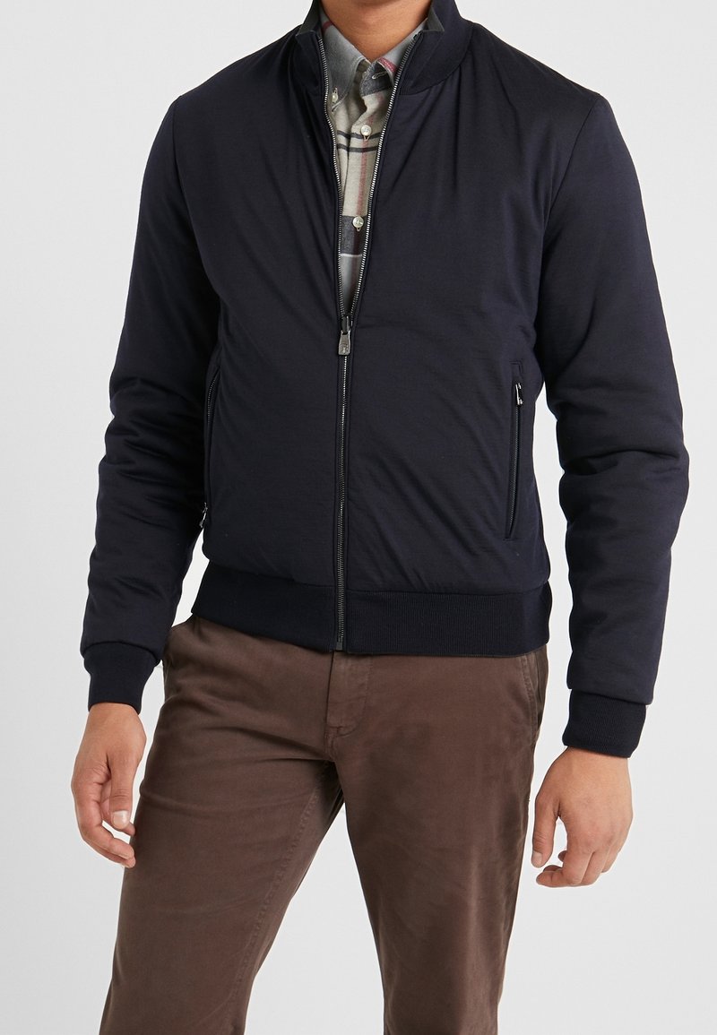 Giubbotto bomber blu scuro con zip frontale, polsini e orlo a coste. Presenta tasche laterali e una texture morbida, indossato sopra una camicia a quadri.
