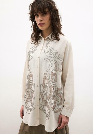 Nő krémszínű, gombos ingben, melyen absztrakt barna és fekete gyöngyminták vannak, lazán viselt barna szoknyával, egyszínű háttér előtt áll.