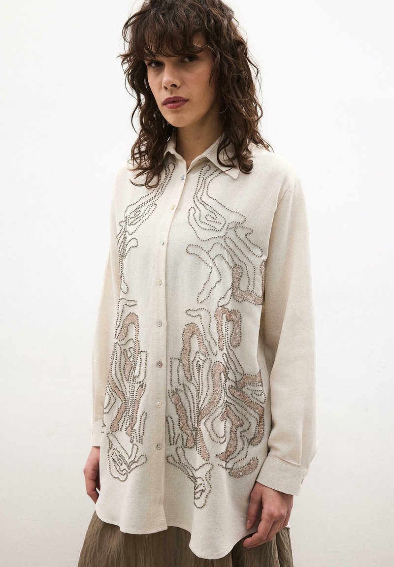 Γυναίκα που φοράει κρεμ πουκάμισο με κουμπιά, διακοσμημένο με αφηρημένα καφέ και μαύρα μοτίβα από χάντρες, συνδυασμένο με φαρδιά καφέ φούστα, στέκεται μπροστά σε απλό φόντο.