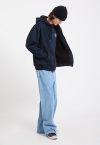 Jeune personne portant un bonnet noir, une veste à capuche bleu marine avec doublure polaire, un jean ample bleu clair et des baskets noires, regardant vers le bas.