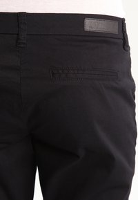 Black cotton pants featuring a back pocket with a horizontal opening, a smooth texture, and a label sewn into the waistband.