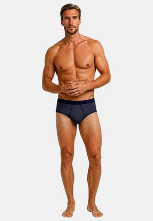 Herren-Slips in Marineblau mit Streifen und einem einfarbigen Bund. Das Material wirkt glatt; die taillierte Passform betont die Körperform. Modell steht barfuß mit verschränkten Armen.