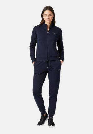 Donna in piedi che indossa una giacca da ginnastica blu navy con zip e pantaloni della tuta coordinati con scarpe da ginnastica nere su sfondo bianco.