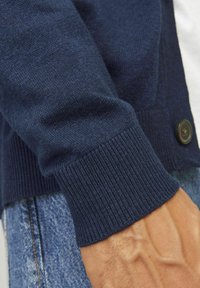 Cardigan blu navy con polsini a coste, texture morbida e un singolo bottone scuro sul davanti. Primo piano di manica e mano.