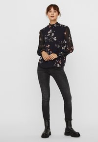 Blusa floreale blu navy con maniche trasparenti, colletto arricciato, e jeans scuri abbinati a stivaletti neri. Presenta motivi pastello sul tessuto.