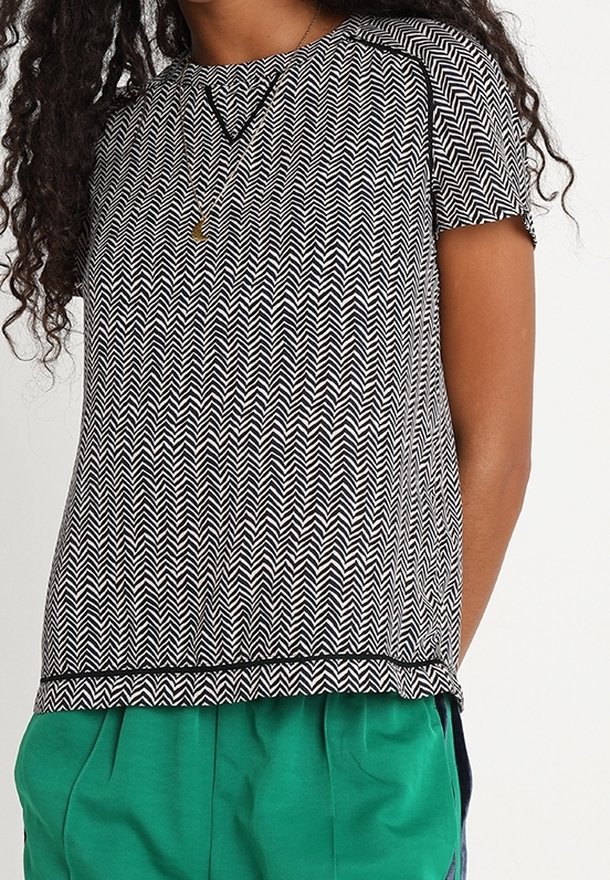 Top de manga corta con un patrón en zigzag en blanco y negro, con un escote redondeado y un ajuste holgado, combinado con pantalones verdes. Textura de tela suave visible.