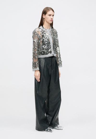 Διαφανές cropped μπουφάν με ασημένιες παγιέτες και μακριά μανίκια, συνδυασμένο με μαύρο δερμάτινο παντελόνι φαρδιάς γραμμής με ψηλή μέση και μεταλλικά μυτερά παπούτσια.