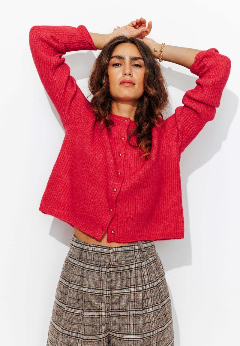 Cardigan en maille côtelée rouge avec cinq boutons, coupe cropped, associé à un pantalon à carreaux gris taille haute avec des rayures verticales.