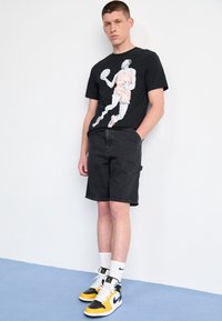 Ung man som står mot en vit vägg, iklädd en svart T-shirt med en grafisk bild av en basketspelare, svarta shorts, vita Nike-strumpor och gula-svarta sneakers.
