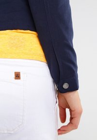 Mörkblå jacka med knappmanschetter, lagd över en gul skjorta. I botten syns vita denimbyxor med en läderetikett.