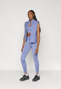 Chaleco sin mangas de color púrpura claro sobre un sujetador deportivo y leggings a juego. Presenta una textura acolchada y el logo de Nike, combinado con zapatillas negras.