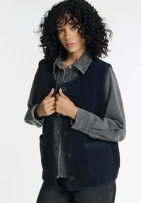 Marineblaue Fleeceweste mit strukturiertem Oberflächen, vorderer Knopfleiste und Seitentaschen. Getragen über einem grauen Denimhemd, was einen geschichteten Look zeigt.