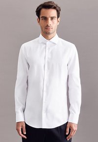 Camicia bianca a maniche lunghe realizzata in tessuto liscio, con colletto classico, chiusura frontale a bottoni e orlo arrotondato, mostrata su uno sfondo neutro.
