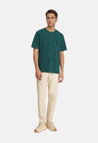 Tyrkysové tričko s krátkým rukávem a texturovaným vzorem, s kapsou na levé části hrudi, v kombinaci s světle béžovými kalhotami a béžovými teniskami.