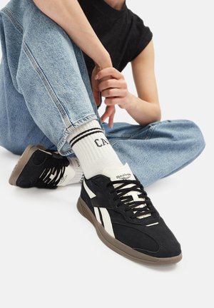 Melni un balti sporta apavi ar teksturētu virsotni, ar šņorēm, gumijas zoli un logotipu uz mēles, apvienoti ar ziliem džinsiem un svītrainām zeķēm.