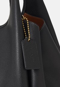 Zwarte gestructureerde leren handtas met een zwart leren label bevestigd aan een gouden kralenketting.