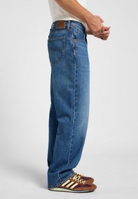 Jeans de mezclilla azul con corte recto, costuras visibles, diseño de cinco bolsillos y una etiqueta de cuero marrón que muestra el logotipo de la marca en la cintura.