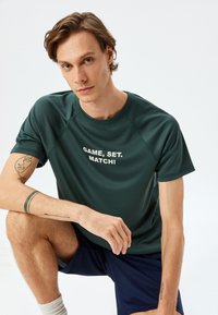 Mörkgrön sporttröja med rund halsringning, korta ärmar och texten "GAME, SET, MATCH!" i vitt. Mjuk, slät textur.