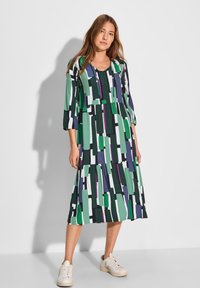 Midi-mekko, jossa on geometrinen printti vihreänä, purppurana ja valkoisena. Mekossa on kolmivartiset hihat ja kerroksellinen hameen muotoilu.