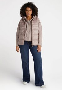 Vaaleanpunainen tilkkutäkkipuffertivesti hupulla, joka on puettu harmaan paidan päälle, yhdistettynä tummansinisii flare-farkkuihin ja valkoisiin kenkiin.