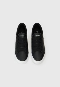 Crne tenisice s glatkim kožnim gornjim dijelom, okruglim prstom i bijelim gumeni potplat. Imaju crne vezice i 'Lacoste' branding na jeziku.