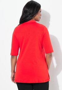 Rode top met korte mouwen en een ronde halslijn, voorzien van een gladde textuur en zijsplitten voor extra comfort. Achteraan zijn er geen extra details.