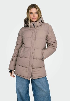 Femme aux cheveux blonds portant une veste matelassée mauve à capuche et un jean large bleu, debout avec une main dans la poche.