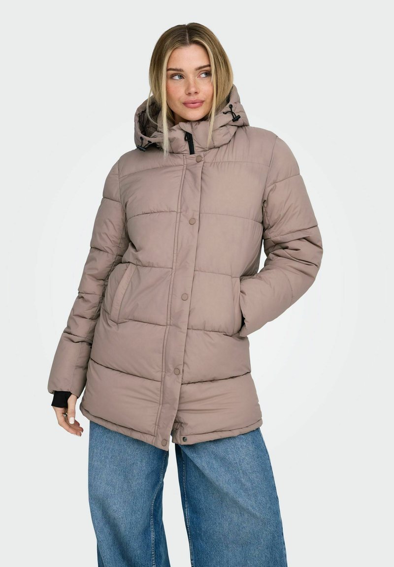 Femme aux cheveux blonds portant une veste matelassée mauve à capuche et un jean large bleu, debout avec une main dans la poche.