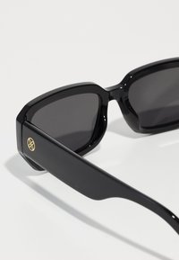 Black sunglasses with a thick, angular frame. Dark tinted lenses and a smooth texture. Gold logo accent on the side.