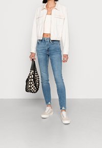 Kvinna iklädd vit kort jacka, ljusbeige kort topp, blå smala jeans, beige sneakers, som håller en svart nätväska med cremefärgad insida.
