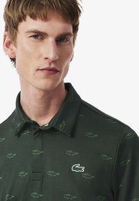 Grön polo t-shirt med subtila krokodilsmönster. Har en knäppt krage och en liten broderad logotyp på bröstet. Slät textur.