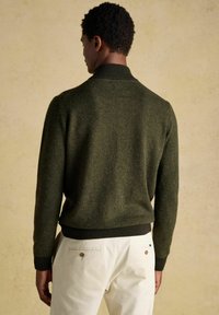 Lühikeste lokkis juustega mees, kes kannab tumeroheline ribatunne salli ja valgeid pükse, nähtuna selja tagant lihtsa tausta ees.